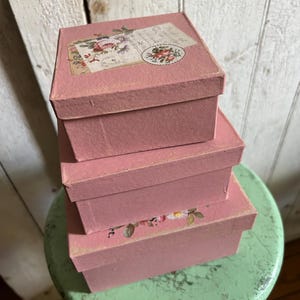 May include: A stack of three square pink boxes with lids. The top box features a vintage-style floral graphic with roses and script text. The bottom box displays a small floral illustration on its lid. Each box has a matte finish and square edges.