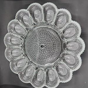 May include: A clear glass deviled egg serving platter featuring a central circular section with a hobnail texture surrounded by twelve individual oval-shaped indentations, each with a ribbed pattern and a textured base for holding eggs.