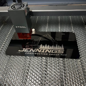 以下が含まれることがあります： 「JENNINGS CONSTRUCTION GROUP」の文字とショベルカーのグラフィックが入った黒い長方形の看板。XTOOLマシンでレーザー彫刻されています。背景はハニカム模様です。