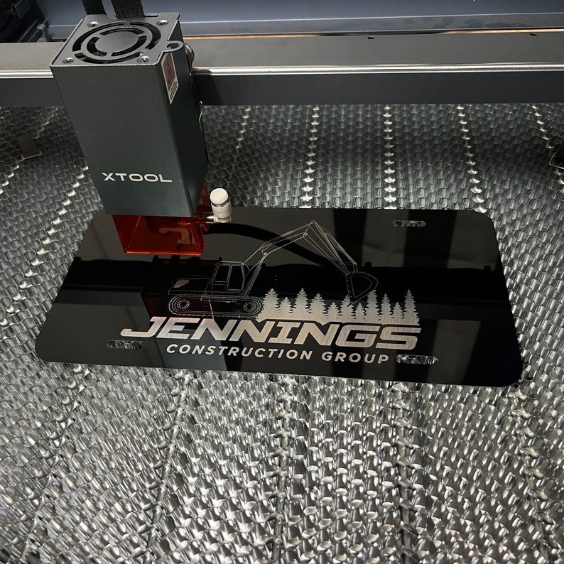 以下が含まれることがあります： 「JENNINGS CONSTRUCTION GROUP」の文字とショベルカーのグラフィックが入った黒い長方形の看板。XTOOLマシンでレーザー彫刻されています。背景はハニカム模様です。