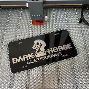 May include: Black license plate with the text "DARK HORSE LASER ENGRAVING" and a knight chess piece logo. The plate is being engraved by an XTOOL laser engraving machine. The background is a silver, textured surface.