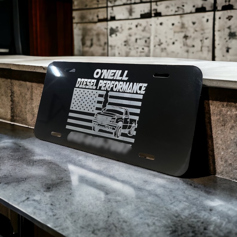 以下が含まれることがあります： 「O'NEILL DIESEL PERFORMANCE」の文字とトラックとアメリカ国旗のデザインが描かれた黒いナンバープレート。ナンバープレートには4つの取り付け穴があり、灰色の表面に表示されています。