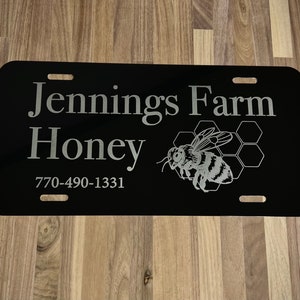 以下が含まれることがあります： 「Jennings Farm Honey」の文字と蜂のグラフィックが入った黒いナンバープレート。電話番号770-490-1331も表示されています。プレートには4つの取り付け穴があります。