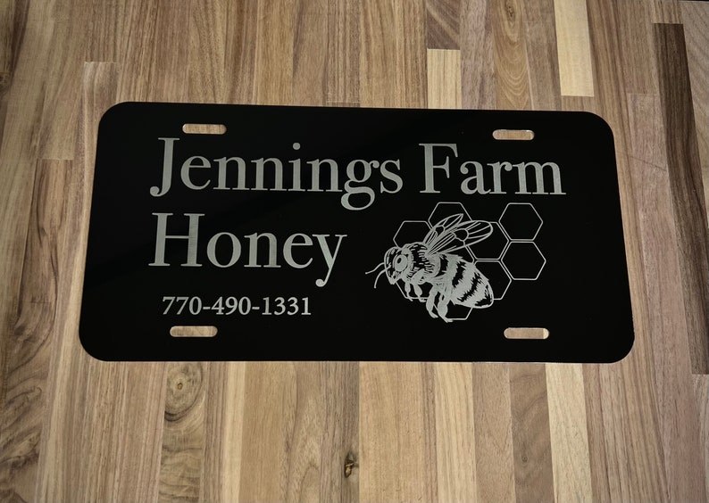 以下が含まれることがあります： 「Jennings Farm Honey」の文字と蜂のグラフィックが入った黒いナンバープレート。電話番号770-490-1331も表示されています。プレートには4つの取り付け穴があります。