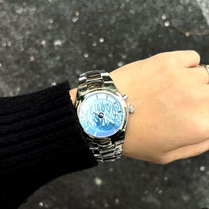 May include: A silver-toned analog wristwatch featuring a metallic link band and a round face. The watch dial displays a vibrant blue and white flame pattern with silver hands and markers, encased in a polished silver-colored metal housing.