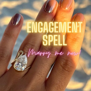 Può includere: Un anello di fidanzamento con diamante a forma di pera incastonato in una fascia d'oro con un design a cigno. L'anello è sul dito di una persona con il testo "ENGAGEMENT SPELL Marry me now!" sovrapposto all'immagine.