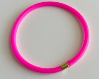 May include: A circular bracelet in a vibrant neon pink color. The bracelet features a smooth, matte-textured band that forms a continuous loop, interrupted only by a single, polished gold-toned cylindrical metal connector piece.