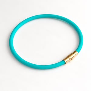May include: A circular turquoise necklace featuring a smooth, flexible cord design. The necklace is secured with a polished gold-tone cylindrical magnetic clasp that connects the two ends of the vibrant, solid-coloured band into a continuous loop.