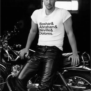 Puede incluir: Imagen en blanco y negro de una persona con una camiseta blanca con el texto "Bashar & Abraham & Neville & Dolores" y pantalones de cuero negro, sentado en una motocicleta.