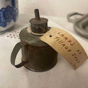 May include: A small, antique oil lamp with a handle and a spout. The lamp is made of aged metal with a rusty appearance. A tag attached to the lamp reads "LAMPS OF VILLAGE INDIA".