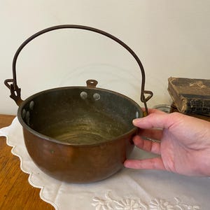 May include: A vintage copper cauldron with a curved handle. The round pot has a dark patina and is riveted. The handle is made of dark metal. The cauldron is sitting on a white, scalloped-edge cloth.