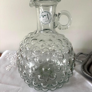 May include: Clear glass pitcher with a round, textured body and a handle. The pitcher has a spout and a label that reads "Made in Mexico". The body of the pitcher is covered in small, bubble-like protrusions.