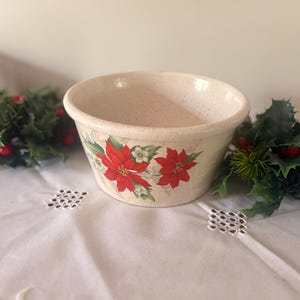 May include: A cream-colored ceramic bowl with a speckled finish and a decorative pattern of red poinsettia flowers and green holly leaves around the exterior. The bowl has a wide, rounded rim and a tapered base, featuring a classic kitchen design.