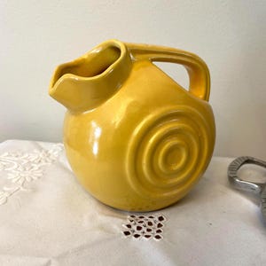 May include: A yellow ceramic pitcher with a rounded body and a handle. The pitcher has a spiral design on the front and a spout for pouring. The pitcher is sitting on a white cloth.
