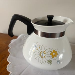 May include: White vintage coffee pot with a black handle and lid, and a silver band. The pot features a floral design with yellow, white, and green flowers. A classic kitchenware item.
