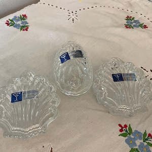 May include: Three clear glass decorative pieces. One is oval-shaped, and the other two are shell-shaped. All three pieces have a textured design and are displayed on a white embroidered tablecloth.