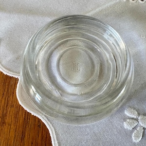 May include: A clear, circular glass coaster with concentric rings. The coaster has a slightly raised edge and a smooth, transparent surface. The coaster is sitting on a white, textured surface.