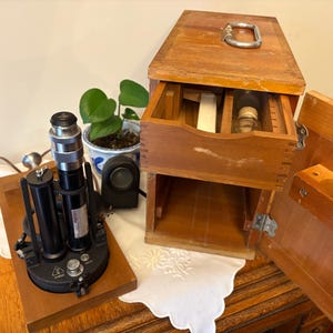 May include: An antique microscope with a black and silver body sits on a wooden base. A wooden box with a handle and open drawer is next to it, revealing various items. A small potted plant is also visible.