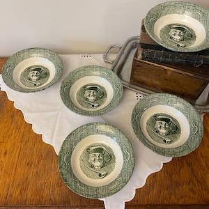 Puede incluir: Cinco cuencos de cerámica verdes y blancos con un retrato de un hombre con sombrero y pipa. Los cuencos están dispuestos sobre un paño blanco, con una bandeja de metal y una caja de madera en el fondo. Los cuencos tienen un borde decorativo.