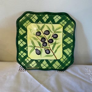 May include: Square ceramic plate with a green plaid border and a central design of an olive branch with black olives. The background of the central design is yellow. The plate has a scalloped edge.