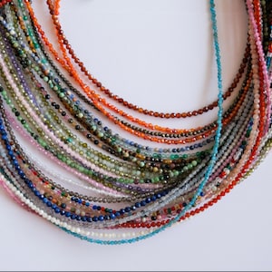 May include: A collection of colourful gemstone beads strung together in a long strand. The beads are various shapes and sizes, and the strand is made up of many different colours, including red, orange, yellow, green, blue, purple, and white.