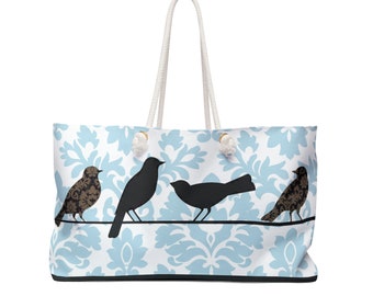 Könnte beinhalten: Eine weiße Tragetasche mit blau-weißem Damastmuster und drei schwarzen Silhouetten von Vögeln, die auf einem Draht sitzen. Die Tasche hat weiße Kordelgriffe und goldfarbene Metalldetails.