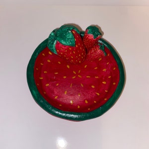 Könnte beinhalten: Eine handbemalte Keramikschale in Form einer Erdbeere. Die Schale ist rot mit gelben Sprenkeln und einem grünen Rand. Drei Erdbeeren mit grünen Blättern sind an der Schale befestigt.