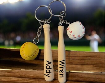 May include: Two baseball-themed keychains. One keychain features a yellow softball charm and a wooden bat charm with the name "Addy". The other keychain has a white baseball charm and a wooden bat charm with the name "Wren".