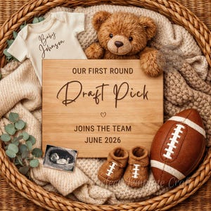 May include: A baby announcement with a wooden sign reading "OUR FIRST ROUND Draft Pick JOINS THE TEAM JUNE 2026". The image includes a football, baby shoes, a teddy bear, a onesie, and an ultrasound photo, all in a woven basket.