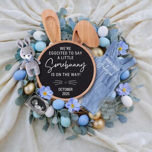 May include: A blue and white baby onesie with the text "Baby Boy" and a name. A wooden letter board with the text "We're eggcited to say a little Somebunny is on the way!" and the date "October 2025". The letter board is surrounded by blue and yellow eggs, blue flowers, and greenery.