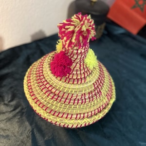 May include: A woven basket with a round, conical shape and a decorative top. The basket is made of natural materials and has a pink and yellow striped pattern. The top of the basket is adorned with several pink and yellow pom-poms.