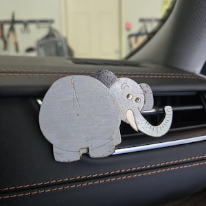 May include: A gray elephant-shaped car air vent clip featuring a cartoon-style design with a large rounded body, a trunk extending to the right, and a small tail. The elephant has visible eyes, a tusk, and textured ear details in a light gray finish.