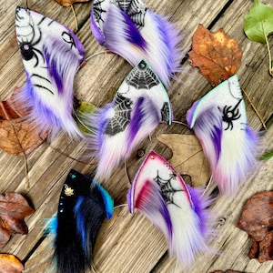 May include: A collection of handmade Halloween-themed cat ear headbands. The ears feature a variety of designs, including spiderwebs, spiders, and colorful fur in shades of purple, white, black, and blue. The headbands are set against a wooden background with autumn leaves.