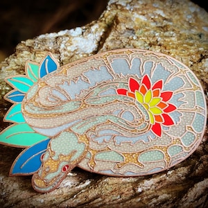 May include: A gold-toned enamel pin featuring a stylized snake with a flower design. The snake is curled up with its head resting on its body. The flower is red, yellow, and orange and is located on the snake's back. The pin has a green and blue background.