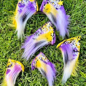 May include: A collection of five triangular faux fur ear accessories featuring a vibrant color palette of yellow, purple, black, and white. Each piece displays a speckled pattern with long, flowing synthetic fur fibers and a small yellow star.