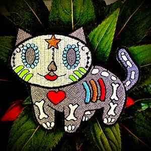 May include: Embroidered iron-on patch of a gray cat with a sugar skull face, rainbow skeleton, and a red heart. The cat has a star on its head and green, blue, and white details on its face.