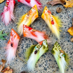 May include: A collection of faux fur animal ears in various colors, including red, orange, yellow, and green, with autumn leaf and acorn accents. Each ear is attached to a thin, gold-colored headband.