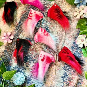May include: A collection of colorful, faux fur cat ears on gold headbands. The ears are in shades of pink, red, and black, some with heart and swirl designs. The ears are surrounded by flowers and greenery.