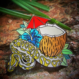 May include: A colorful enamel pin featuring a coconut with a red umbrella, a blue hibiscus flower, and a yellow and white snake.