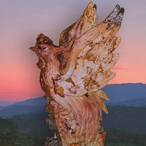 May include: A detailed sculpture of a rooster with outstretched wings, carved from stone. The rooster features shades of brown, beige, and white, with intricate feather detailing. The sculpture is set against a backdrop of a sunset over a mountain range.