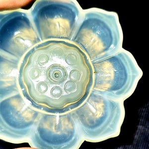 Puede incluir: Primer plano de un objeto de vidrio con forma de flor con un diseño central de círculos y un borde exterior en forma de pétalos. El vidrio tiene una calidad translúcida y opalescente con tonos de azul, blanco y amarillo. El objeto está sostenido por una mano.
