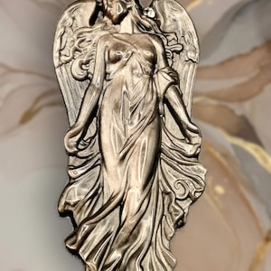 May include: A metallic bronze-toned relief sculpture of a standing angel with large, detailed wings. The figure is depicted with flowing robes that drape down to the feet, with arms slightly extended at the sides and a serene, downward-facing head.