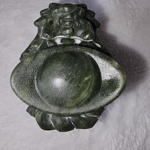 May include: A carved, dark green stone sculpture with a circular, bowl-like shape. The sculpture has a textured surface and appears to be a decorative item. The design includes stylized floral or leaf elements.