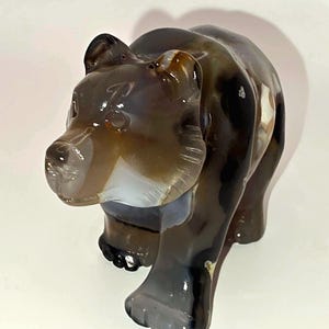 May include: A carved bear figurine made of polished brown and translucent white agate stone. The sculpture features a smooth, glossy surface with natural banding and dark inclusions, depicting a bear in a walking stance with visible facial details.