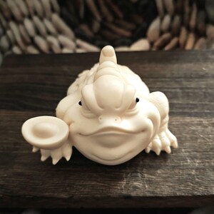 May include: A cream-colored, smiling frog figurine holding a coin. The frog has detailed features, including textured skin and small black eyes. The figurine is placed on a dark wooden surface.