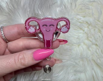 May include: Pink glitter uterus charm with a smiling face, attached to a clear plastic retractable badge holder.