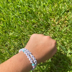 Puede incluir: Dos pulseras de cuentas con cuentas de letras blancas que deletrean "TEAM CONRATSITP". Las pulseras tienen cuentas de colores pastel entre las cuentas de letras. Las pulseras están en una muñeca sobre un fondo de hierba verde.