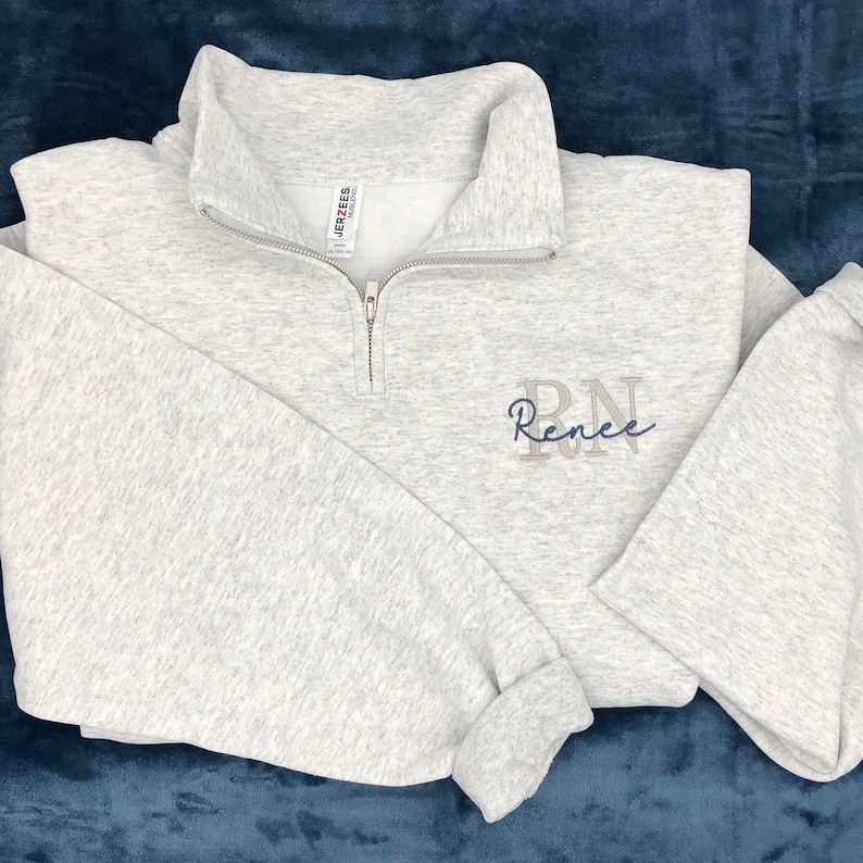 Ash gray 1/4 quarter zip sweatshirt with custom embroidery featuring RN in light gray in all caps with name Renee in cursive font in smokey blue thread.