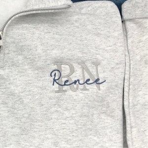 Close up of embroidery for Ash gray 1/4 quarter zip sweatshirt with custom embroidery featuring RN in light gray in all caps with name Renee in cursive font in smokey blue thread.