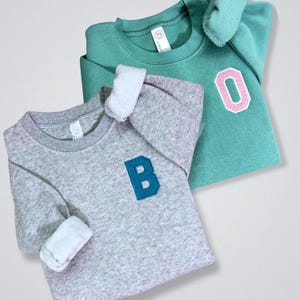 May include: Two crew neck sweatshirts. One is heather gray with a teal "B" patch, the other is teal with a pink-outlined "0" patch. Both have white cuffs and a small tag on the neckline.